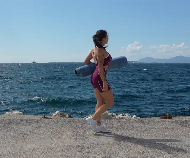 Femme en tenue de sport marchant sur une jetée rocailleuse, portant un tapis de yoga sous le bras, avec la mer et des montagnes à l'horizon.