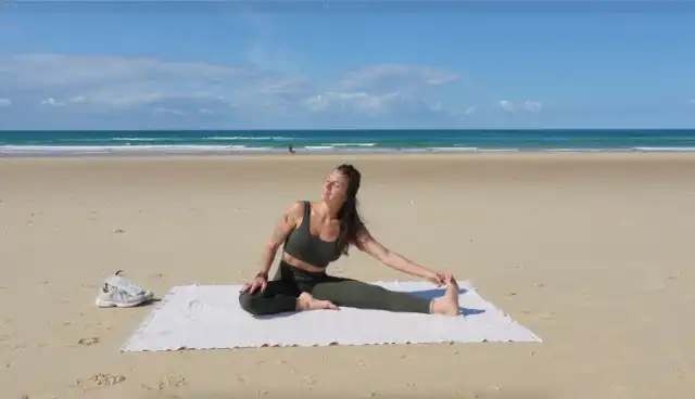 Femme en tenue de sport verte effectuant une posture de yoga Janu Sirsasana sur une serviette blanche à la plage.