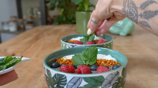 Une main dépose une feuille de menthe sur un bol rempli de fruits rouges, myrtilles, pollen et copeaux de noix de coco.