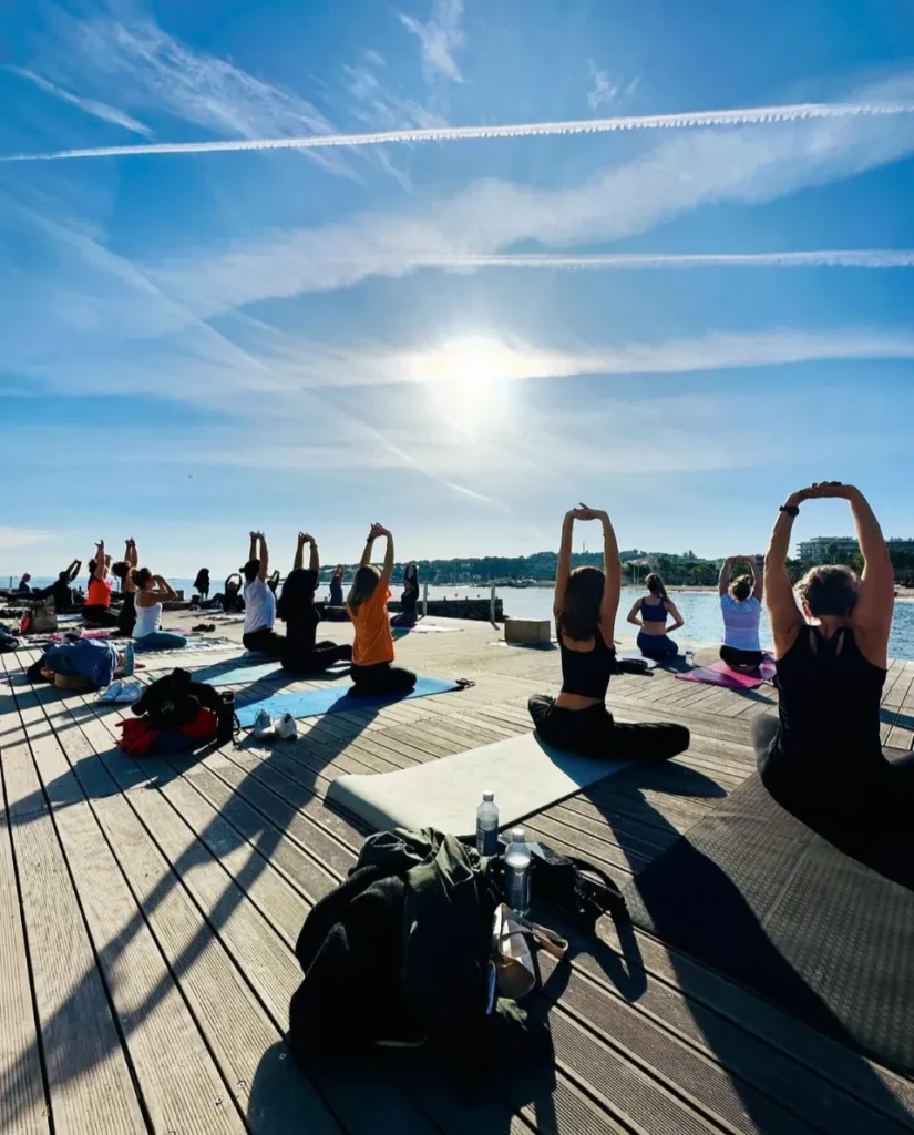Un groupe de personnes pratique le yoga en plein air sur une terrasse en bois au bord de la mer, bras levés vers le ciel ensoleillé et nuageux.