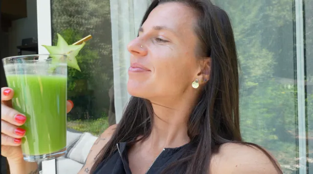 Une femme aux cheveux bruns tient un verre de jus vert vif décoré d'une étoile de carambole.