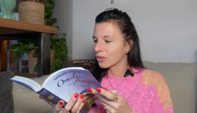 Femme aux ongles rouges, lisant un livre intitulé 'Oracles des Anges' de Doreen Virtue. Elle porte un pull rose et orange.