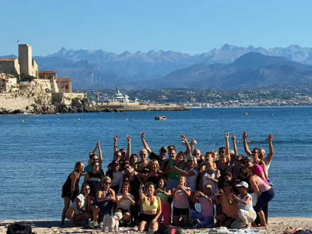 Groupe de personnes souriantes et joyeuses sur la plage, devant la mer et la ville d'Antibes avec les montagnes en arrière-plan.
