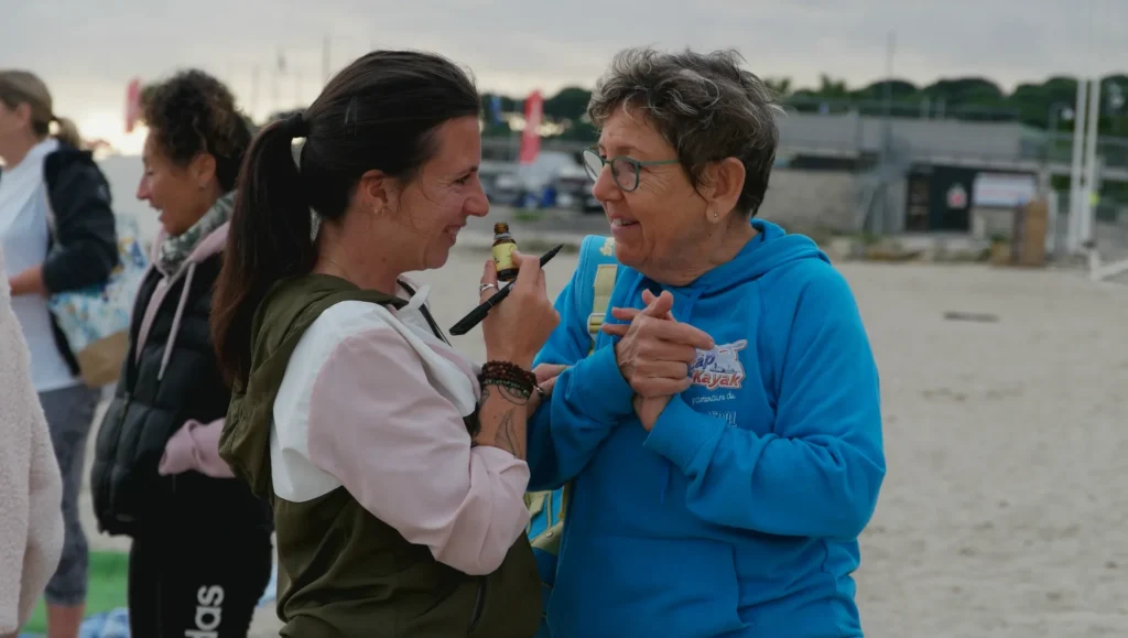 Deux femmes sourient sur la plage, l'une appliquant du maquillage à l'autre avec un petit flacon et un pinceau.