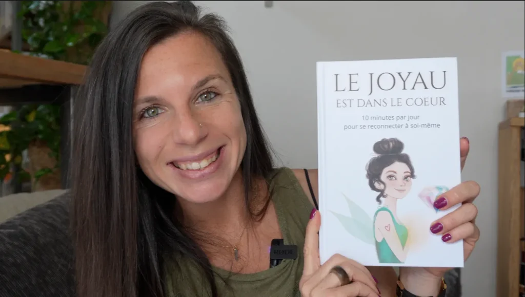 Une femme souriante tient un livre intitulé « Le joyau est dans le cœur », un guide pour se reconnecter à soi-même.
