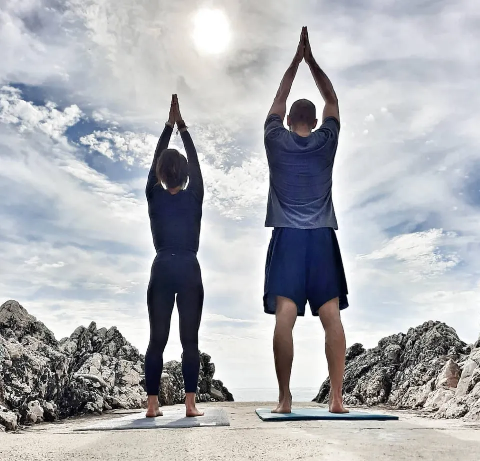 Deux personnes pratiquant le yoga sur des tapis, bras levés et mains jointes, devant un ciel nuageux au coucher du soleil, avec des rochers en arrière-plan.