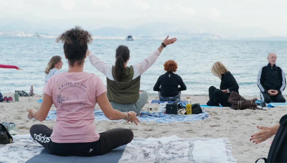 Un groupe de personnes pratique le yoga en tailleur sur une plage, avec la mer en arrière-plan.