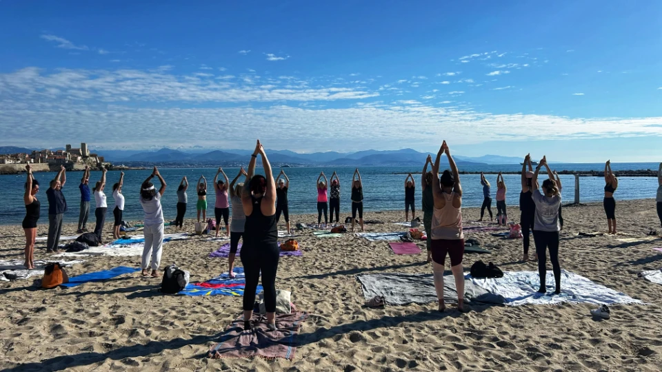 Un groupe de personnes pratique le yoga sur une plage de sable, face à la mer, sous un ciel bleu avec des nuages et un paysage montagneux à l'horizon.