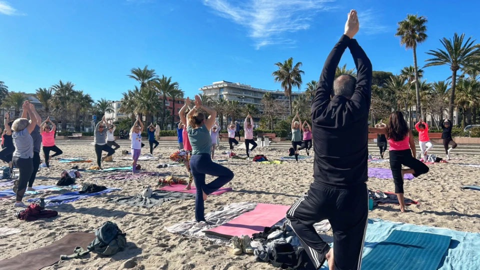 Un groupe de personnes pratiquant le yoga en plein air sur le sable d'une plage, avec des palmiers et des bâtiments en arrière-plan.