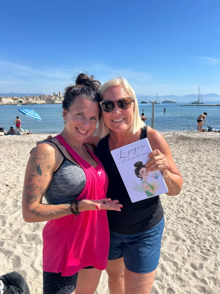 Deux femmes souriantes sur une plage de sable, l'une tenant un livre intitulé "Le joyau est dans le cœur".