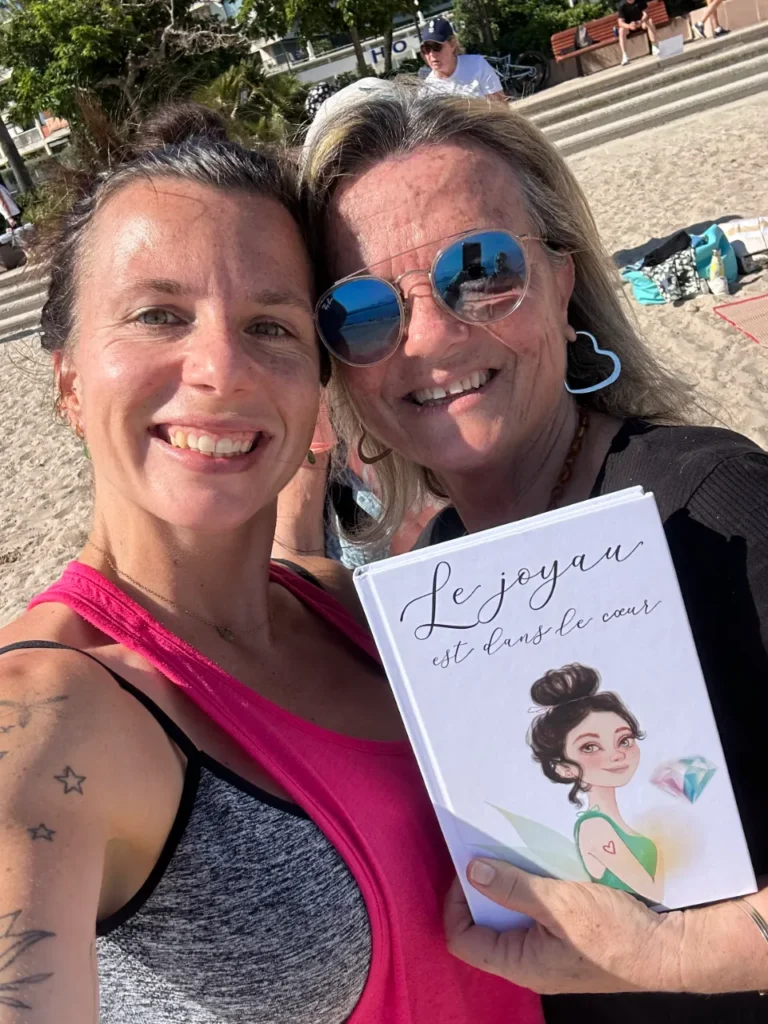Deux femmes souriantes prennent un selfie sur une plage ensoleillée. L'une tient un livre blanc avec une illustration de fée et le texte "Le joyau est dans le cœur".