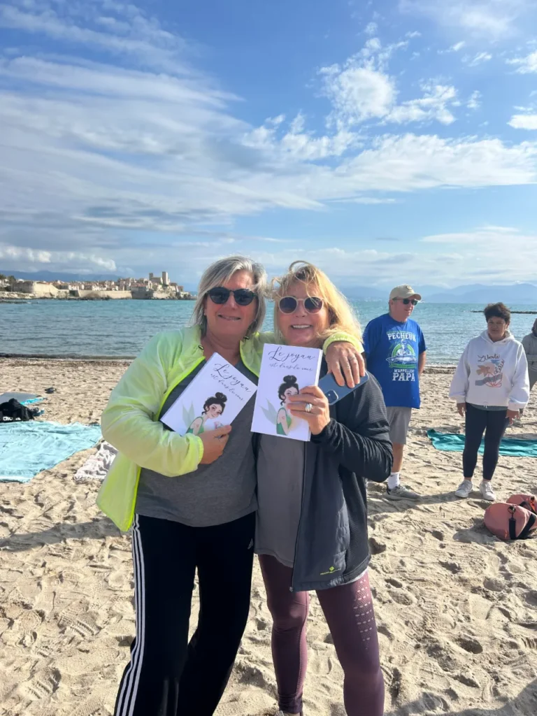Deux femmes souriantes posent sur une plage ensoleillée, tenant chacune un livre illustré sur le yoga, avec une ville au bord de l'eau en arrière-plan.