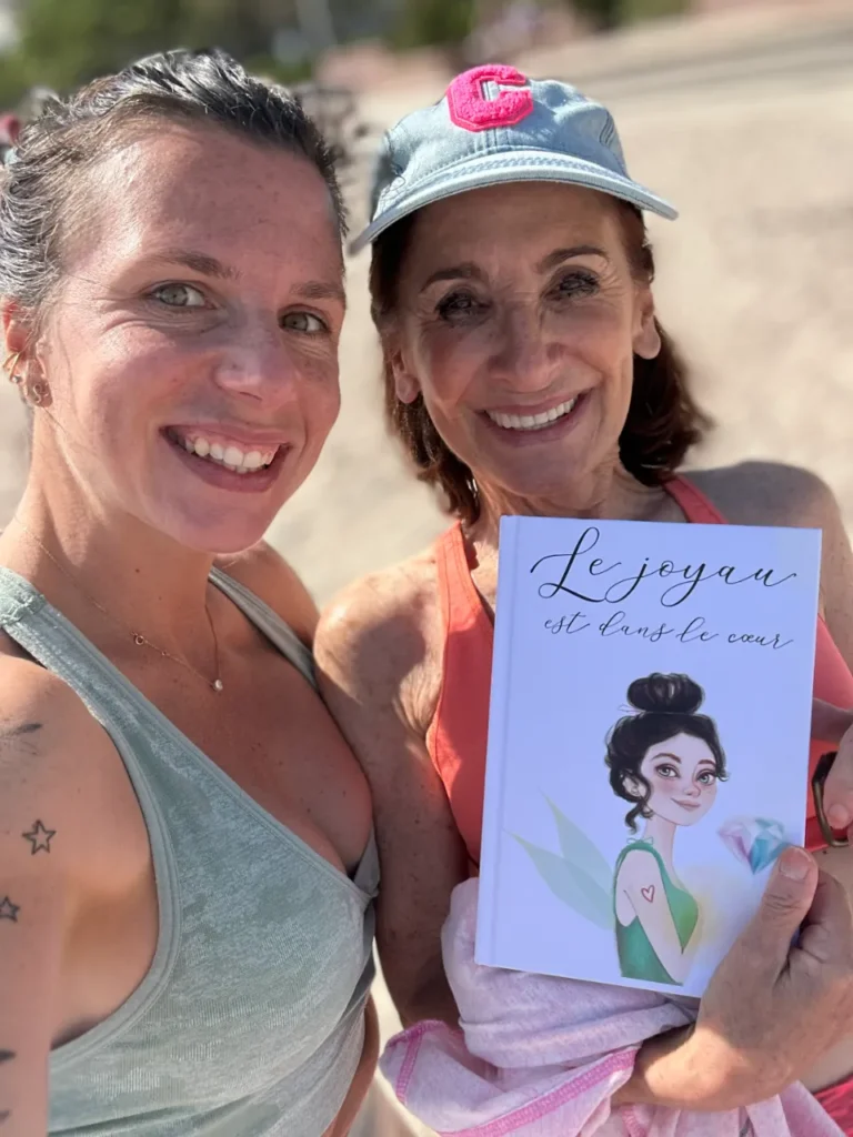 Deux femmes souriantes se tiennent à l'extérieur. La femme de droite tient un livre intitulé "Le joyau est dans le cœur" avec une illustration de fée.
