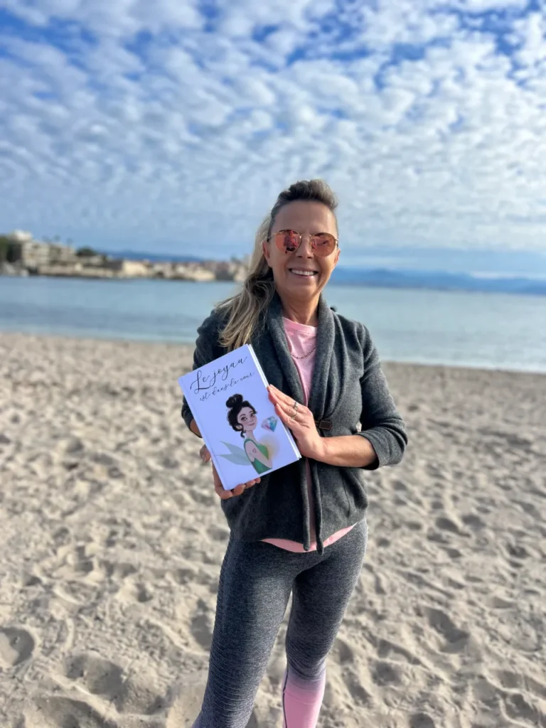 Une femme souriante, le soleil dans les yeux, tenant un livre intitulé 'Le joyau est dans le cœur', avec une illustration de fée sur la couverture.