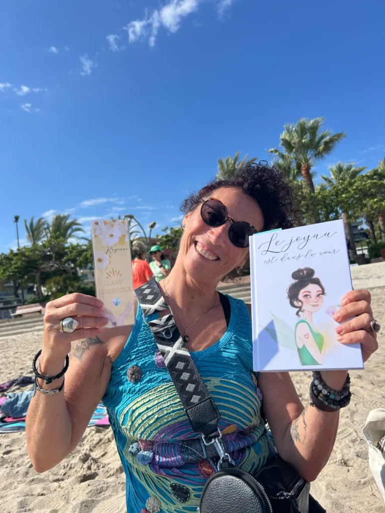 Une femme au sourire éclatant tient un livre intitulé "Le joyau est dans le cœur" et un marque-page "Reyvana Le Joyau Est Dans Le Cœur" sur fond de plage et de cocotiers.