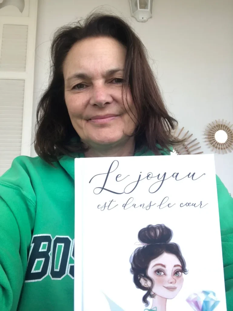 Une femme souriante tenant un livre illustré avec une fille et un diamant, dont le titre est "Le joyau est dans le cœur".