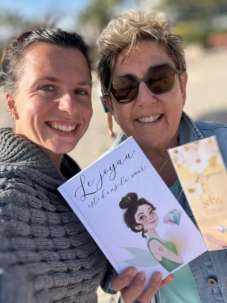 Deux femmes souriantes se prennent en selfie, l'une tenant un livre orné d'une illustration de fée et de diamants, l'autre un prospectus similaire.