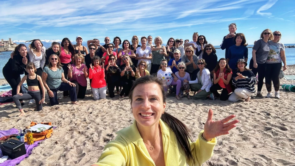 Un groupe de personnes souriantes s'est réuni sur une plage ensoleillée pour une séance de yoga, avec la mer et un ciel bleu en arrière-plan.