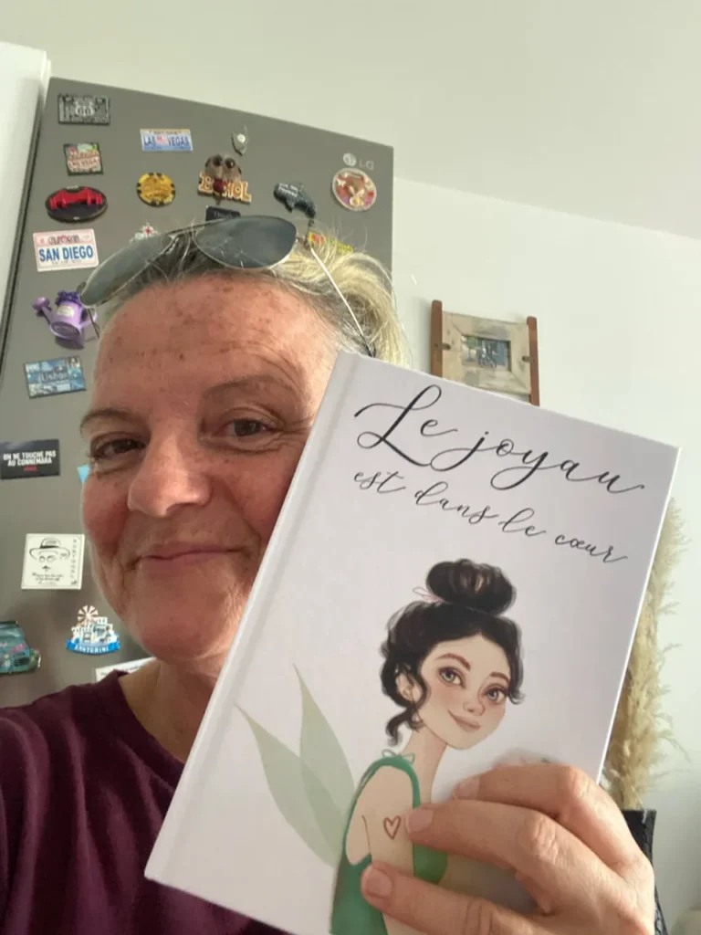 Une femme souriante tient un livre intitulé "Le joyau est dans le cœur", illustré par une fée aux ailes vertes, sur fond de réfrigérateur décoré.
